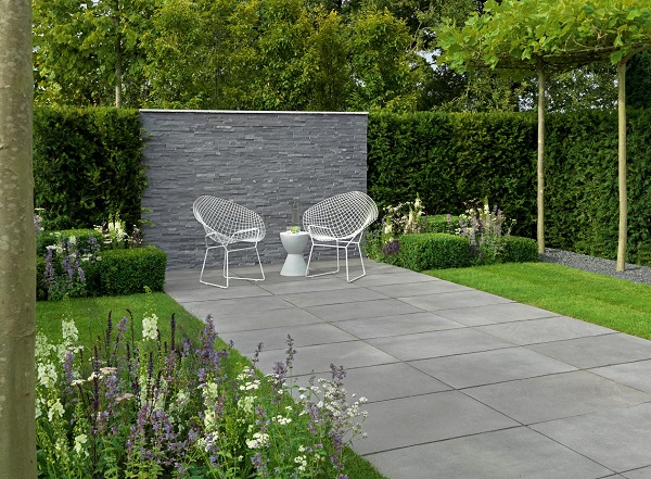 Grey limestone paving with white round outdoor chairs and small white table with grey wall behind with grass, shrubs, flowers and bushes on either side