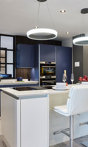 large kitchen with bright circular cieling lights, white bartop with built in cooking top and white high chairs, dark blue cupboards with built in oven and grill and fridge freezer