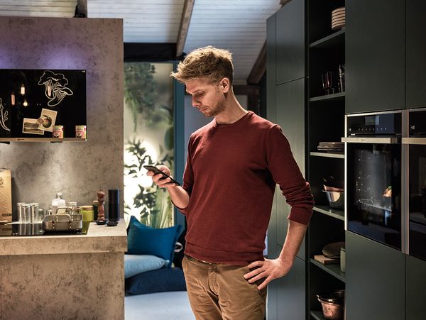 Man standing in kitchen looking at his phone with cooking top to his right and ovens to his left